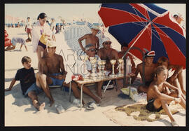 Grupo de pessoas e jovens sentados na areia à sombra de sombrinha de praia, com uma mesa com taças.